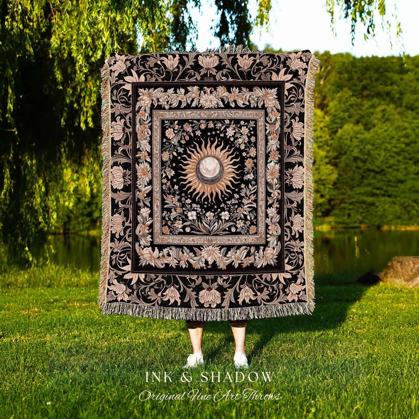 May include: A black woven throw blanket with a gold and white sun and moon design in the center. The design is surrounded by a floral pattern. The blanket has fringe on all sides. The text 'INK & SHADOW Original Fine Art Throws' is at the bottom of the blanket.
