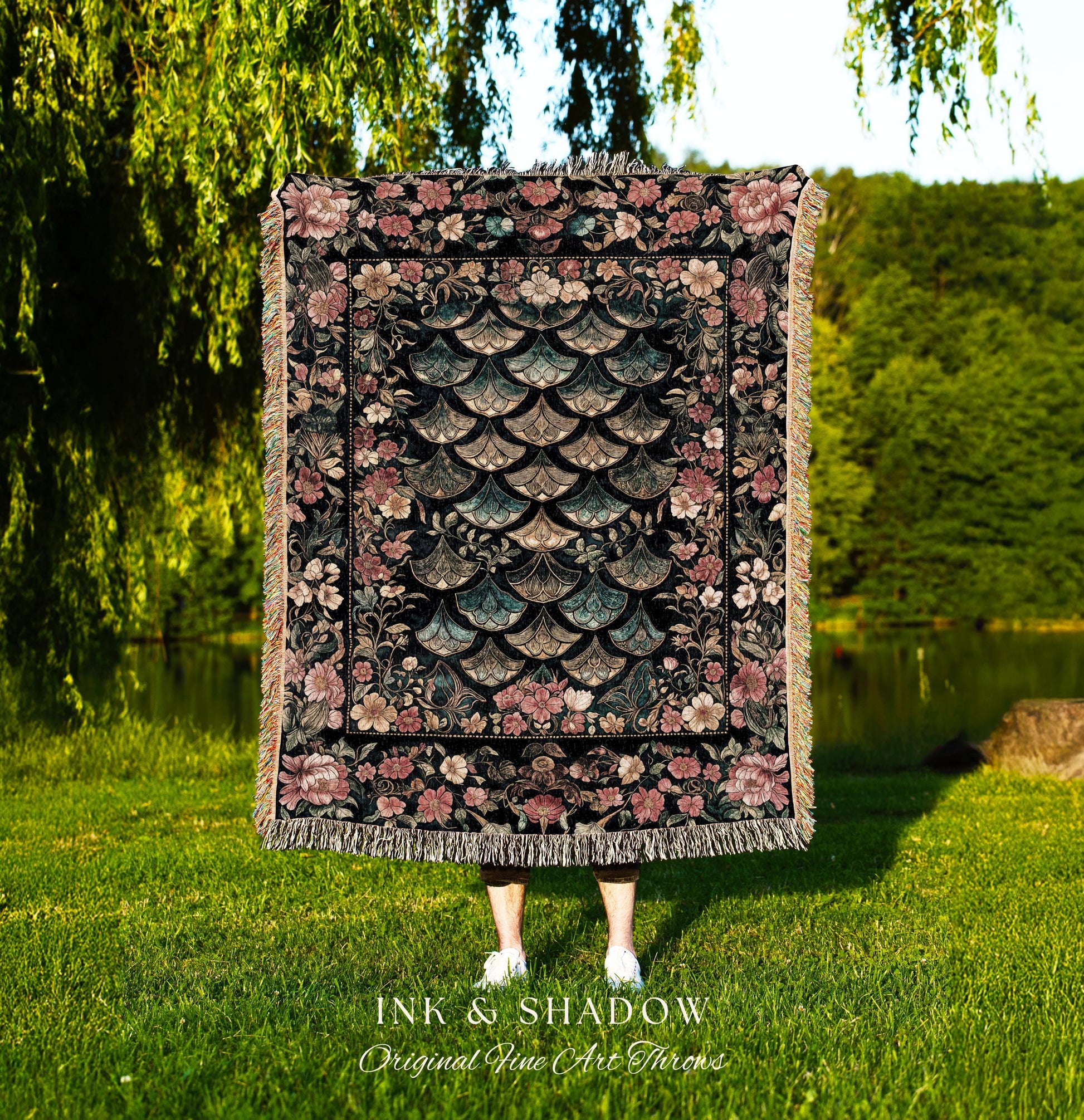 May include: A woven tapestry throw blanket with a black background and a floral border. The center of the blanket features a pattern of overlapping teal and pink scales. The blanket has fringe along the bottom edge. The text &#39;INK & SHADOW Original Fine Art Throws&#39; is printed below the blanket.