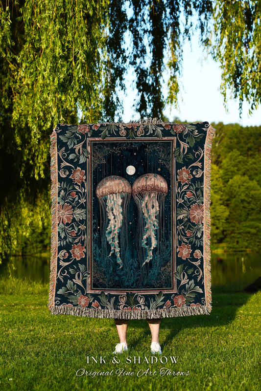 May include: A woven tapestry throw blanket with a dark blue background and a floral border. The center of the blanket features a dark blue and green illustration of two jellyfish swimming in front of a full moon. The text &#39;INK & SHADOW Original Fine Art Throws&#39; is printed at the bottom of the blanket.