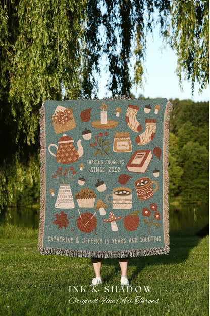 May include: A woven throw blanket with a teal background and a variety of autumn-themed illustrations, including a teapot, a candle, a jar of honey, a book, a cupcake, mushrooms, a mug of hot chocolate, and a ball of yarn. The text on the blanket reads &#39;Sharing Snuggles Since 2008&#39; and &#39;Catherine & Jeffery 15 Years and Counting.&#39; The blanket is made by Ink & Shadow Original Fine Art Throws.