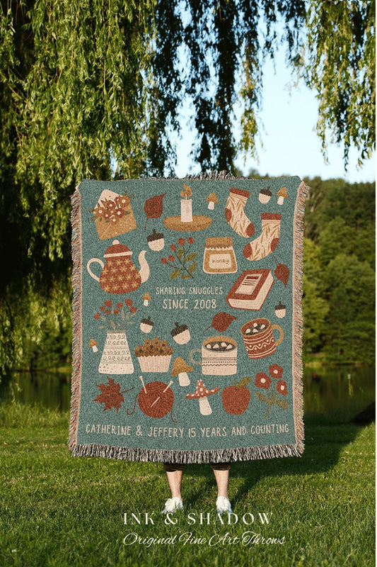 May include: A woven throw blanket with a teal background and a variety of autumn-themed illustrations, including a teapot, a candle, a jar of honey, a book, a cupcake, mushrooms, a mug of hot chocolate, and a ball of yarn. The text on the blanket reads &#39;Sharing Snuggles Since 2008&#39; and &#39;Catherine & Jeffery 15 Years and Counting.&#39; The blanket is made by Ink & Shadow Original Fine Art Throws.