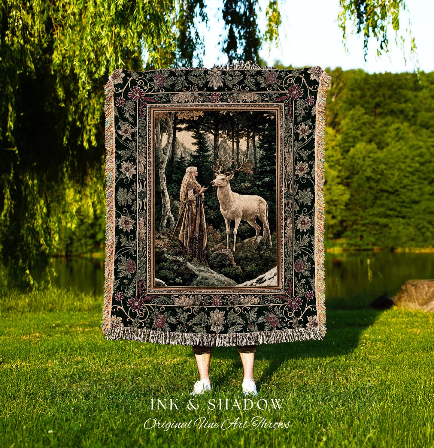 May include: A tapestry throw blanket with a dark green background and a floral border. The center of the tapestry features a woman in a long white dress standing in a forest with a white deer. The tapestry has the words &#39;Ink & Shadow Original Fine Art Throws&#39; at the bottom.