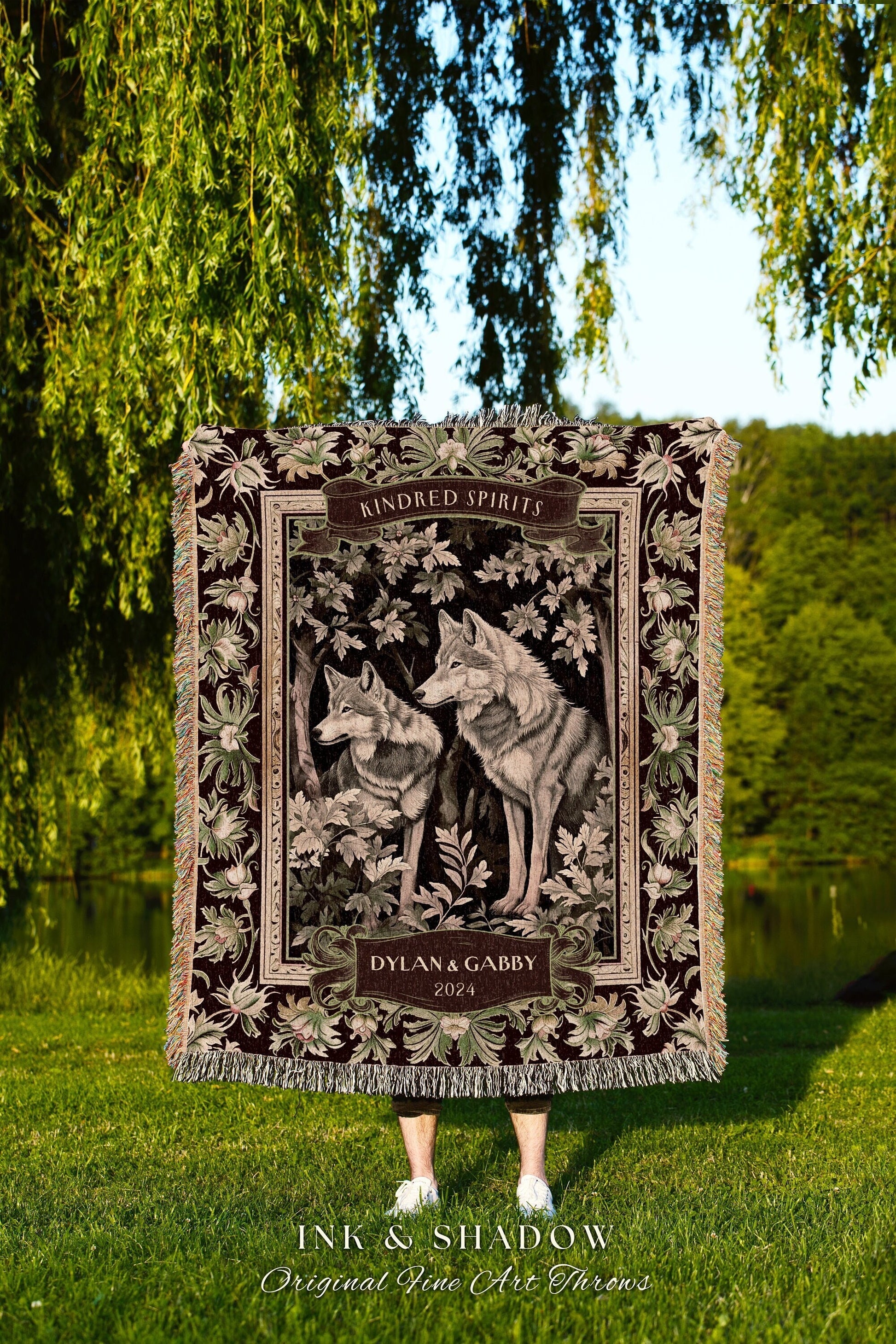 May include: A tapestry throw blanket with a dark brown background. The blanket features a floral border with two gray wolves in the center. The wolves are standing in a forest setting with green leaves. The blanket has a banner at the top that reads &#39;Kindred Spirits&#39; and a banner at the bottom that reads &#39;Dylan & Gabby 2024&#39;. The blanket is made by Ink & Shadow Original Fine Art Throws.