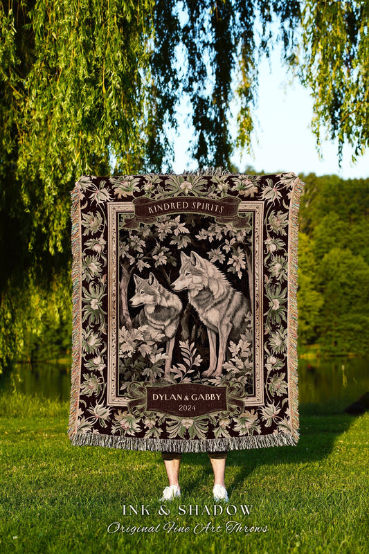 May include: A tapestry throw blanket with a dark brown background. The blanket features a floral border with two gray wolves in the center. The wolves are standing in a forest setting with green leaves. The blanket has a banner at the top that reads &#39;Kindred Spirits&#39; and a banner at the bottom that reads &#39;Dylan & Gabby 2024&#39;. The blanket is made by Ink & Shadow Original Fine Art Throws.