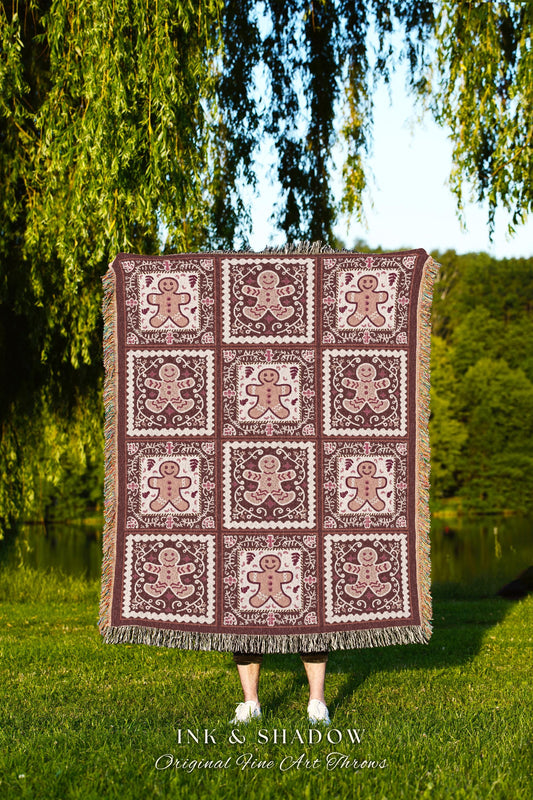 May include: A woven throw blanket with a gingerbread man pattern. The blanket is brown with a white and pink design. The blanket has a fringe border. The text &#39;INK & SHADOW Original Fine Art Throws&#39; is printed on the bottom of the blanket.