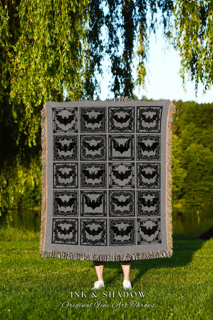 May include: A black and gray woven throw blanket with a repeating pattern of bats in ornate frames. The blanket has a fringe border and the text &#39;INK & SHADOW Original Fine Art Throws&#39; is printed at the bottom.