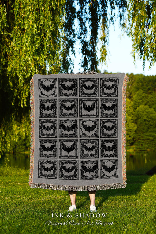 May include: A black and gray woven throw blanket with a repeating pattern of bats in ornate frames. The blanket has a fringe border and the text &#39;INK & SHADOW Original Fine Art Throws&#39; is printed at the bottom.