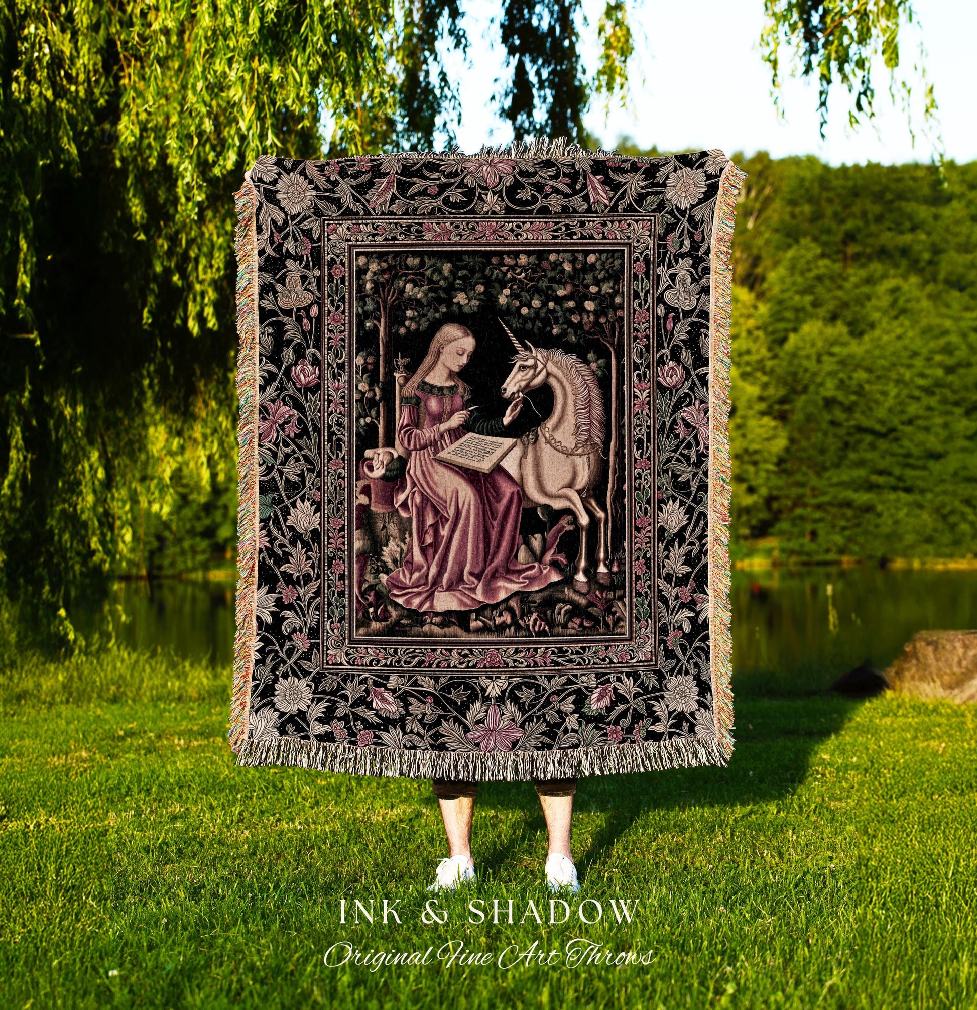 May include: A woven tapestry throw blanket with a black background and a pink and cream floral pattern. The center of the blanket features a framed image of a woman in a pink dress sitting with a unicorn. The text &#39;INK & SHADOW Original Fine Art Throws&#39; is printed below the blanket.