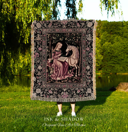 May include: A woven tapestry throw blanket with a black background and a pink and cream floral pattern. The center of the blanket features a framed image of a woman in a pink dress sitting with a unicorn. The text &#39;INK & SHADOW Original Fine Art Throws&#39; is printed below the blanket.
