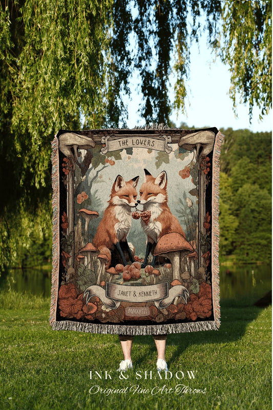 May include: A woven tapestry throw blanket with a black background and a colorful design. The design features two foxes facing each other, surrounded by mushrooms, berries, and vines. The text 'THE LOVERS' is at the top of the design, and the text 'JANET & KENNETH MMXXIII' is at the bottom of the design. The throw blanket is a part of the 'INK & SHADOW Original Fine Art Throws' collection.