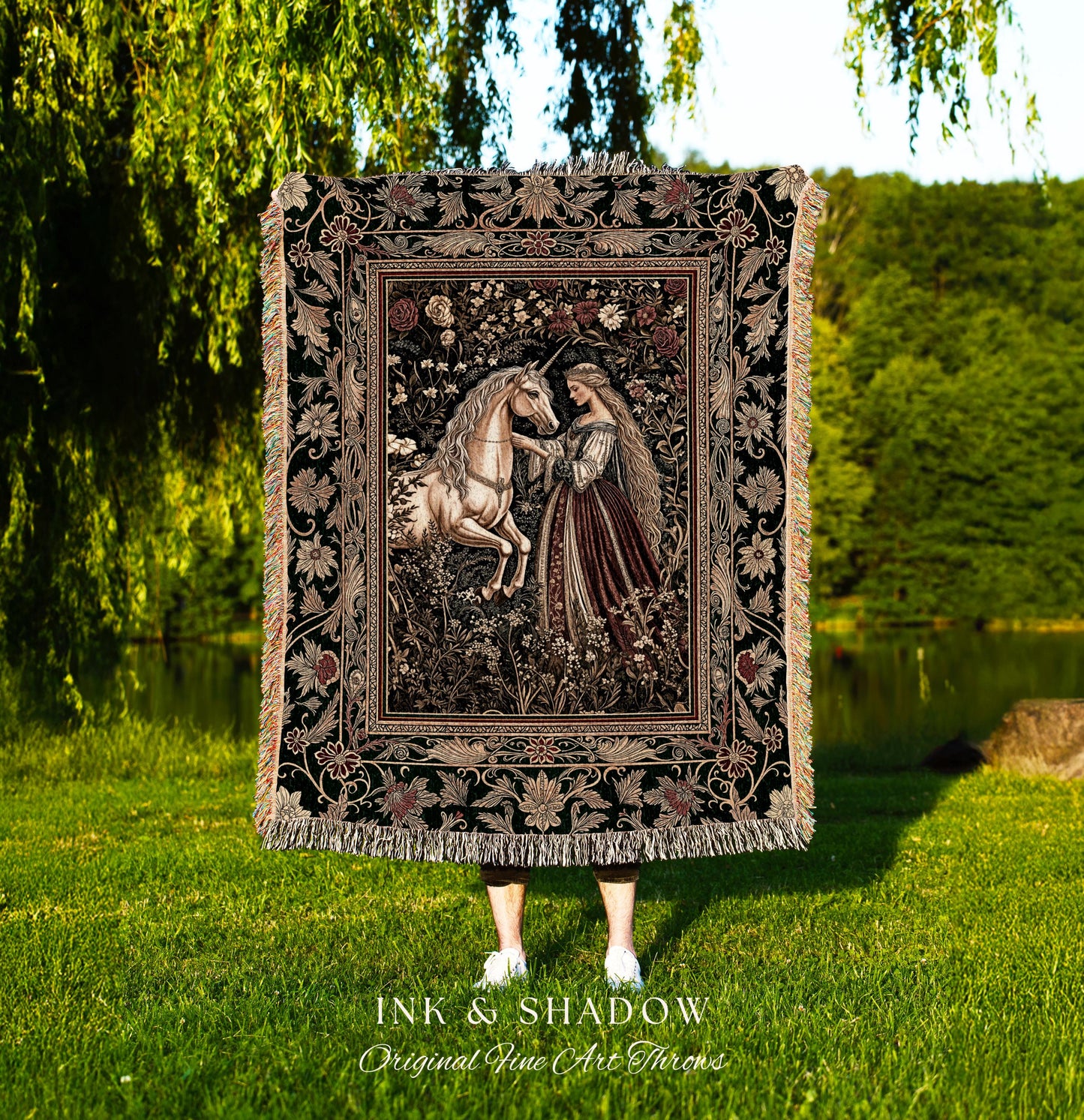 May include: A black woven tapestry throw blanket with a floral border and a central image of a woman in a long gown interacting with a white unicorn in a forest setting. The text &#39;INK & SHADOW Original Fine Art Throws&#39; is printed below the blanket.