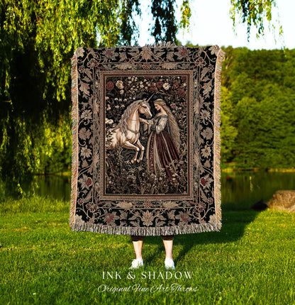 May include: A black woven tapestry throw blanket with a floral border and a central image of a woman in a long gown interacting with a white unicorn in a forest setting. The text &#39;INK & SHADOW Original Fine Art Throws&#39; is printed below the blanket.