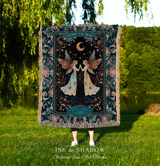 May include: A woven tapestry throw blanket with a dark blue background and a floral pattern. The center of the blanket features a design of two fairies with wings, a crescent moon, and stars. The text &#39;GEMINI - THE TWINS&#39; is at the top of the design. The text &#39;MAY 21 - JUNE 20&#39; is at the bottom of the design. The text &#39;INK & SHADOW Original Fine Art Throws&#39; is at the bottom of the blanket.