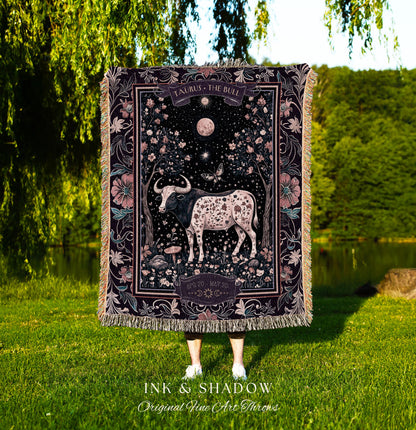 May include: A tapestry throw blanket with a dark background and a pink floral border. The center of the blanket features a black and white bull with pink spots. The text &#39;TAURUS THE BULL&#39; is at the top of the blanket and &#39;APRIL 20 MAY 20&#39; is at the bottom. The text &#39;INK & SHADOW Original Fine Art Throws&#39; is at the bottom of the blanket.