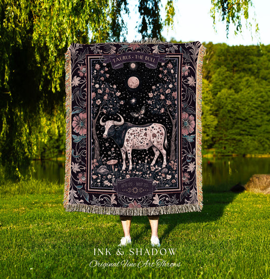 May include: A tapestry throw blanket with a dark background and a pink floral border. The center of the blanket features a black and white bull with pink spots. The text &#39;TAURUS THE BULL&#39; is at the top of the blanket and &#39;APRIL 20 MAY 20&#39; is at the bottom. The text &#39;INK & SHADOW Original Fine Art Throws&#39; is at the bottom of the blanket.