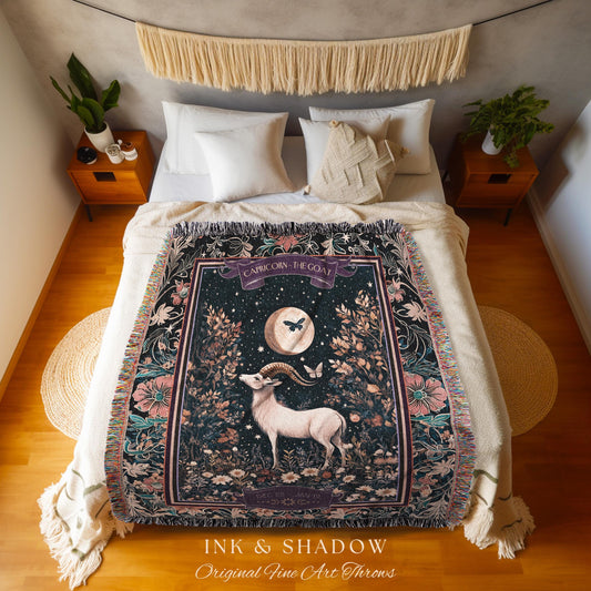 May include: A woven tapestry throw blanket with a dark background and a colorful floral design. The center of the blanket features a black and white illustration of a goat with a crescent moon and stars. The text &#39;Capricorn, The Goat&#39; is printed in a white banner above the goat. The blanket is displayed on a bed with white bedding and a wooden floor. The text &#39;Ink & Shadow Original Fine Art Throws&#39; is printed below the blanket.