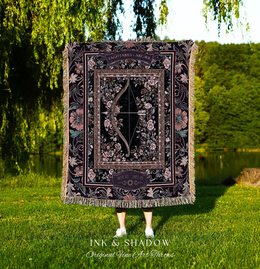 May include: A woven tapestry throw blanket with a black background and a floral pattern. The center of the blanket features a large, ornate frame with the words 'Sagittarius - Archer' and 'Nov 22 - Dec 21' inside. The frame contains a stylized image of a bow and arrow with floral accents. The blanket has a fringed edge.