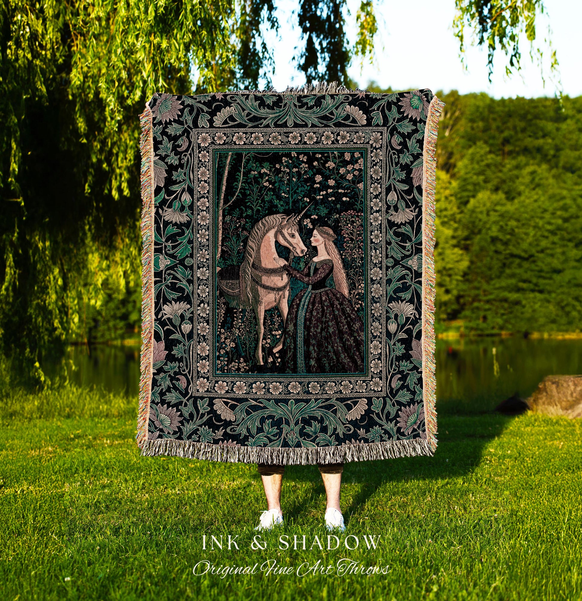 May include: A woven tapestry throw blanket with a black background and a green, pink, and white floral design. The center of the blanket features a scene of a woman in a long dress reaching out to a unicorn. The blanket has fringe on the edges and the words &#39;INK & SHADOW Original Fine Art Throws&#39; are printed at the bottom.