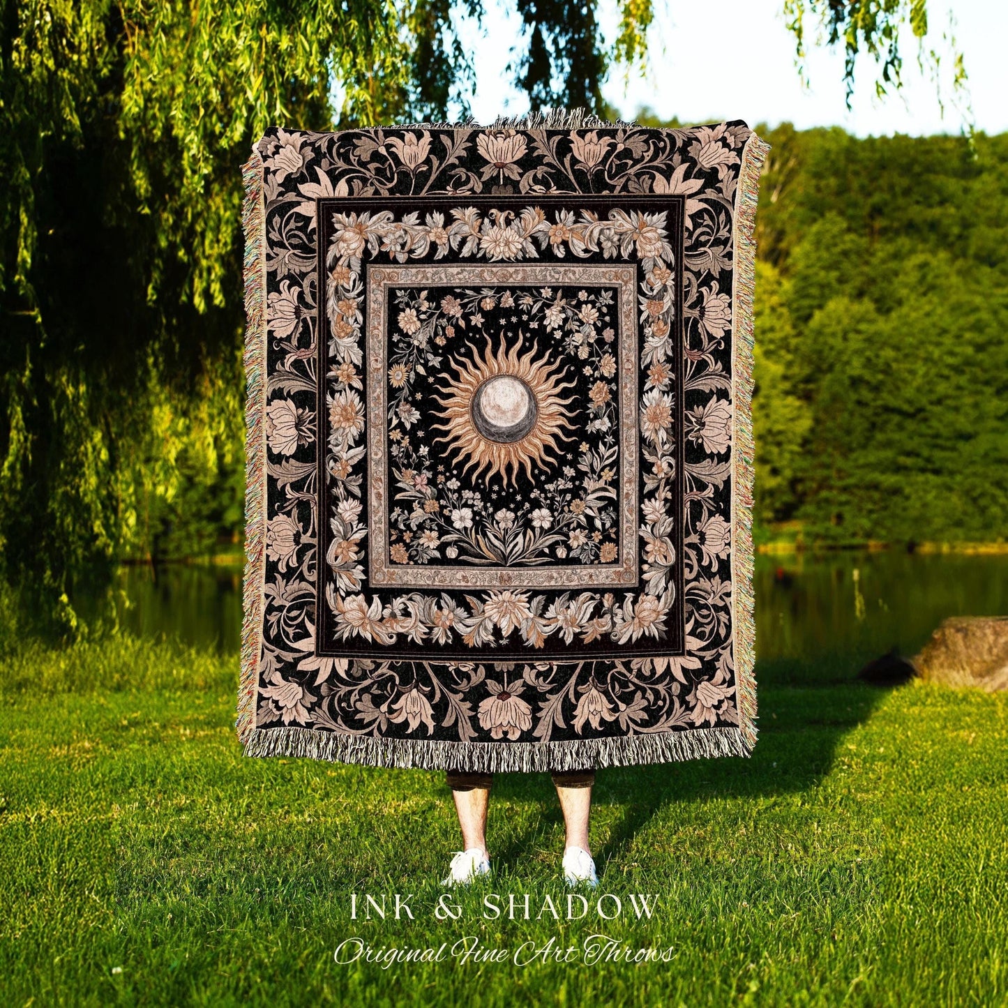 May include: A black woven throw blanket with a gold and white sun and moon design in the center. The design is surrounded by a floral pattern. The blanket has fringe on all sides. The text &#39;INK & SHADOW Original Fine Art Throws&#39; is at the bottom of the blanket.