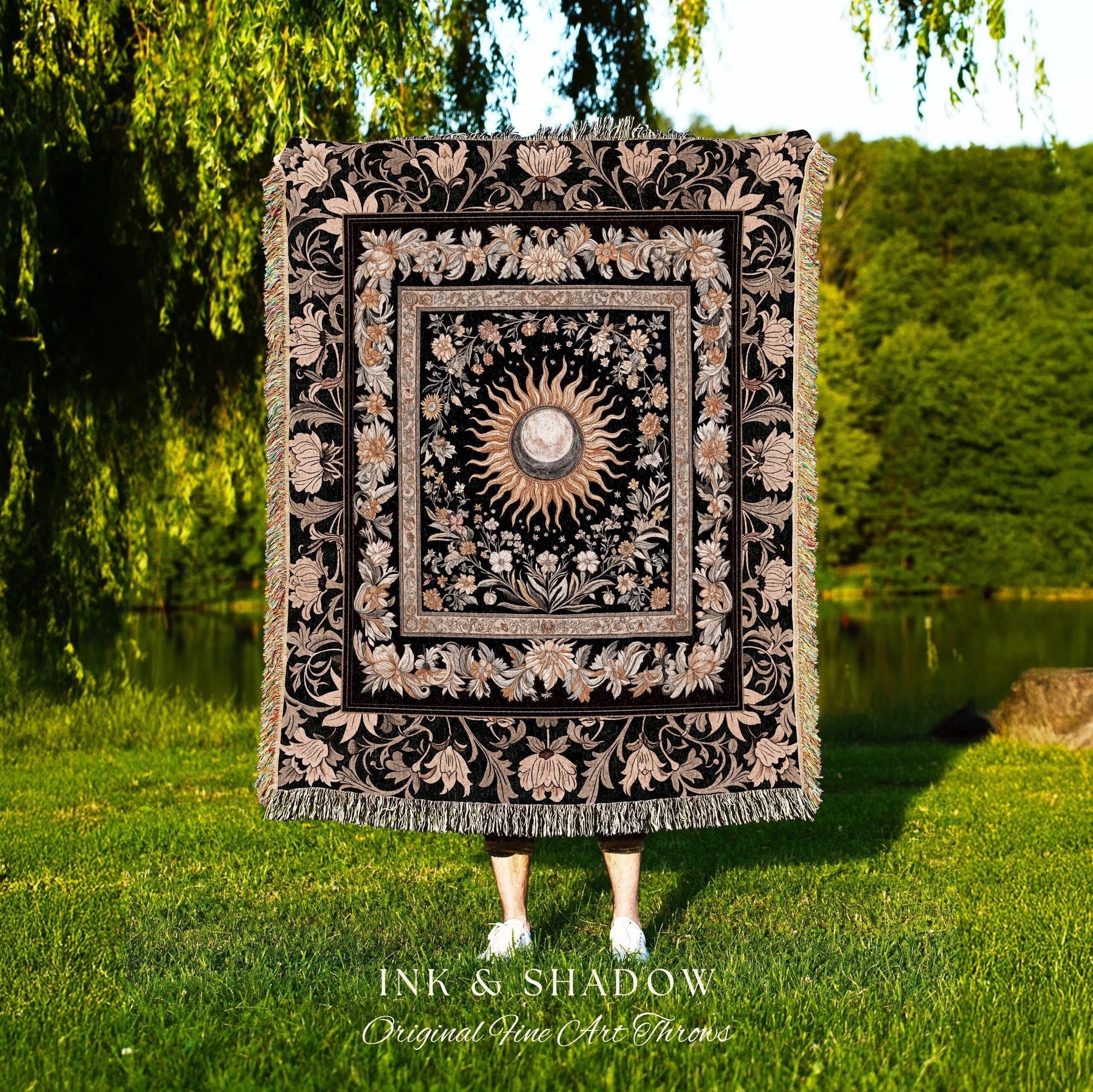 May include: A black woven throw blanket with a gold and white sun and moon design in the center. The design is surrounded by a floral pattern. The blanket has fringe on all sides. The text &#39;INK & SHADOW Original Fine Art Throws&#39; is at the bottom of the blanket.