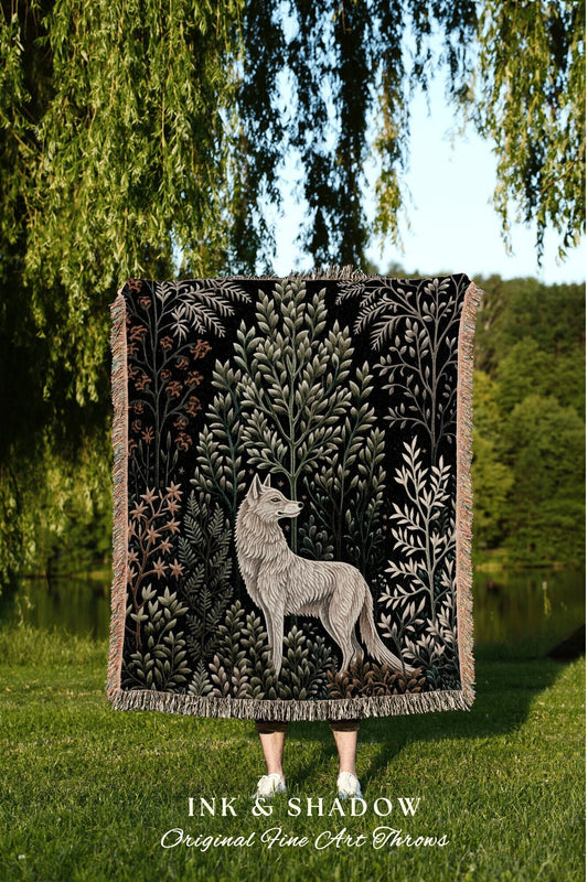 May include: A woven tapestry throw blanket with a black background and a white wolf standing in a forest of green trees. The throw blanket has fringe on all sides. The text &#39;INK & SHADOW Original Fine Art Throws&#39; is printed below the blanket.