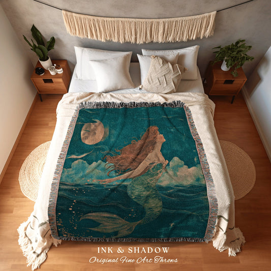 May include: A woven throw blanket with a teal background featuring a mermaid with long brown hair swimming towards a full moon. The throw blanket has a fringe border and the words &#39;INK & SHADOW Original Fine Art Throws&#39; at the bottom.
