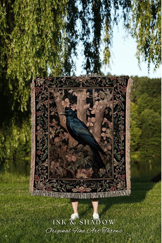 May include: A black woven tapestry throw blanket with a dark blue bird perched on a branch in a forest scene. The throw blanket has a black background with a floral pattern in shades of green, pink, and brown. The throw blanket has a fringe border. The text &#39;INK & SHADOW Original Fine Art Throws&#39; is printed below the blanket.