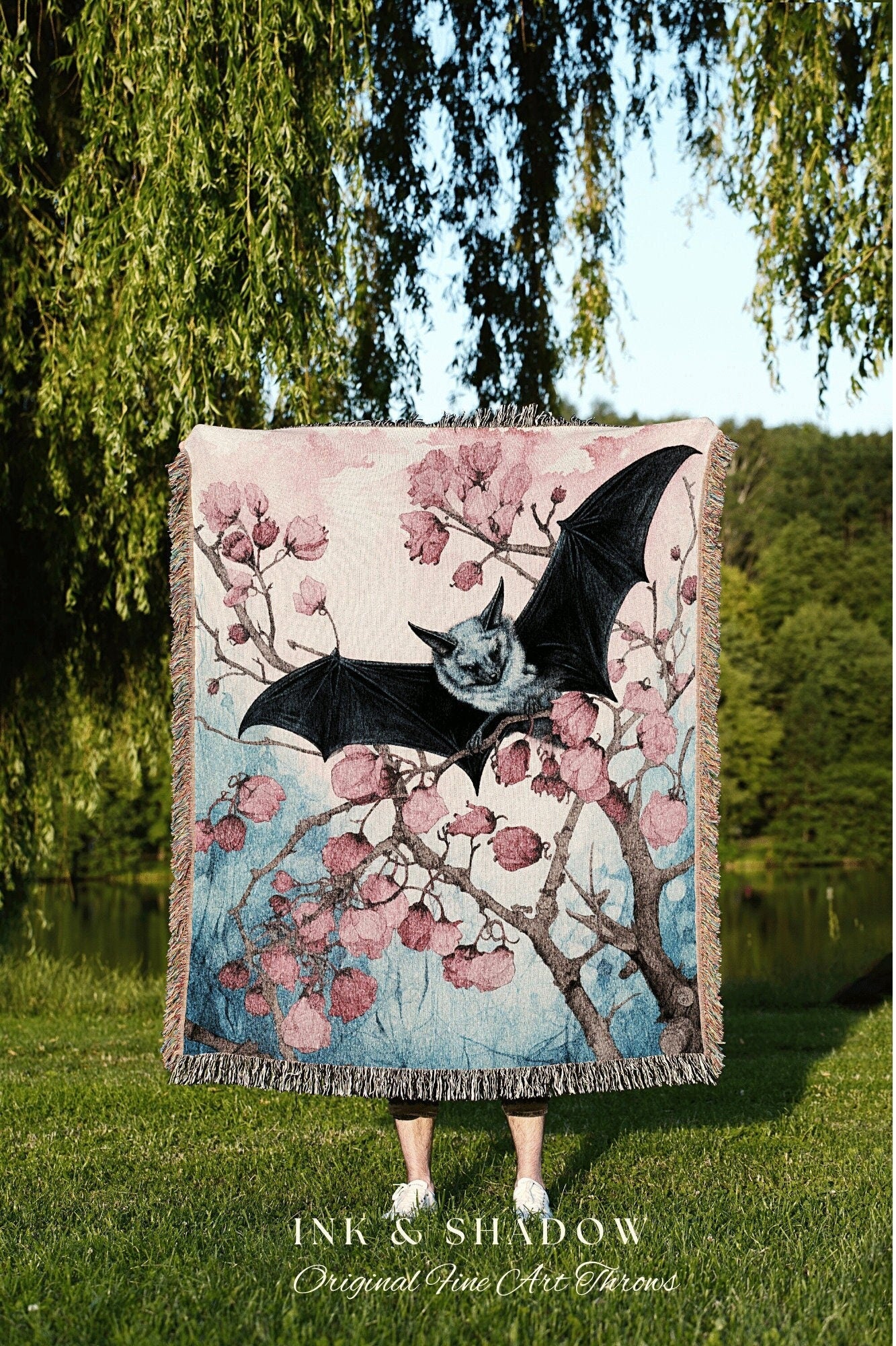 May include: A woven tapestry throw blanket with a pink, blue, and white color scheme. The blanket features a black bat perched on a branch of a flowering tree. The text &#39;INK & SHADOW Original Fine Art Throws&#39; is printed below the blanket.
