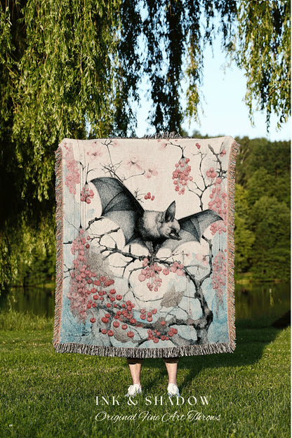 May include: A woven tapestry throw blanket with a pink, blue, and gray color scheme. The blanket features a black bat perched on a branch with pink berries. The text &#39;INK & SHADOW Original Fine Art Throws&#39; is printed at the bottom of the blanket.