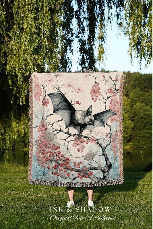 May include: A woven tapestry throw blanket with a pink, blue, and gray color scheme. The blanket features a black bat perched on a branch with pink berries. The text &#39;INK & SHADOW Original Fine Art Throws&#39; is printed at the bottom of the blanket.