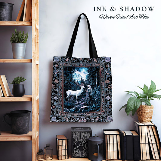 May include: A black tote bag with a woven tapestry design. The tapestry features a white deer and a woman standing in a forest with a mountain in the background. The tote bag has the text &#39;INK & SHADOW Woven Fine Art Tote&#39; printed on the top.