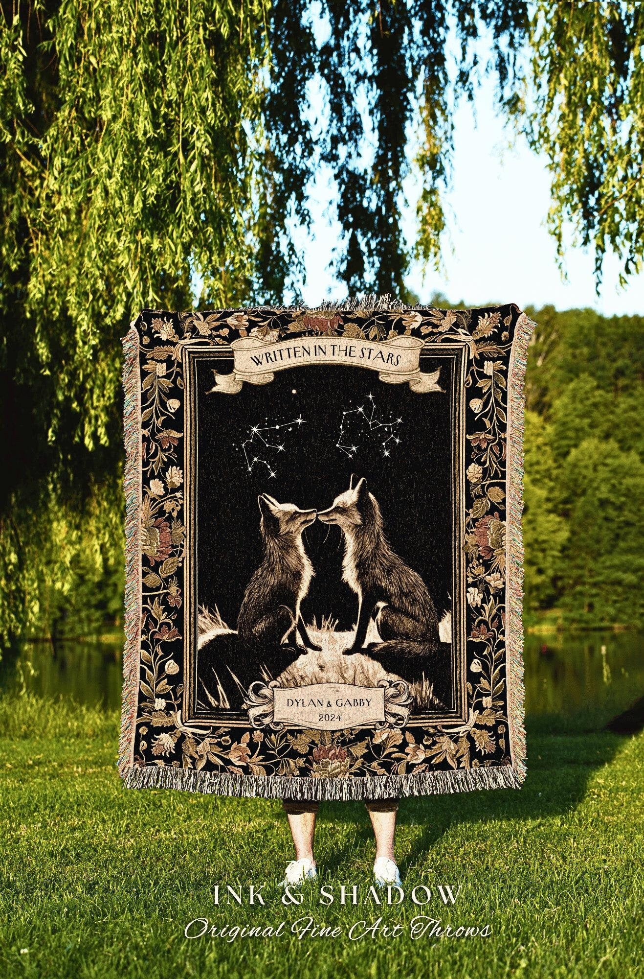 May include: A black tapestry throw blanket with a floral border. The center of the blanket features a constellation of stars with two foxes kissing under the stars. The text &#39;Written in the Stars&#39; is above the foxes and the text &#39;Dylan & Gabby 2024&#39; is below the foxes. The text &#39;Ink & Shadow Original Fine Art Throws&#39; is at the bottom of the blanket.