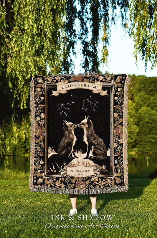 May include: A black tapestry throw blanket with a floral border. The center of the blanket features a constellation of stars with two foxes kissing under the stars. The text &#39;Written in the Stars&#39; is above the foxes and the text &#39;Dylan & Gabby 2024&#39; is below the foxes. The text &#39;Ink & Shadow Original Fine Art Throws&#39; is at the bottom of the blanket.