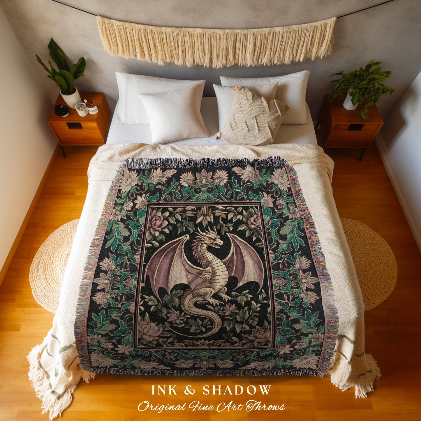 May include: A woven tapestry throw blanket with a black background and a green, pink, and gold floral design. A gold dragon with pink wings is featured in the center of the design. The throw blanket is displayed on a bed with white bedding. The text &#39;INK & SHADOW Original Fine Art Throws&#39; is printed on the bottom of the blanket.