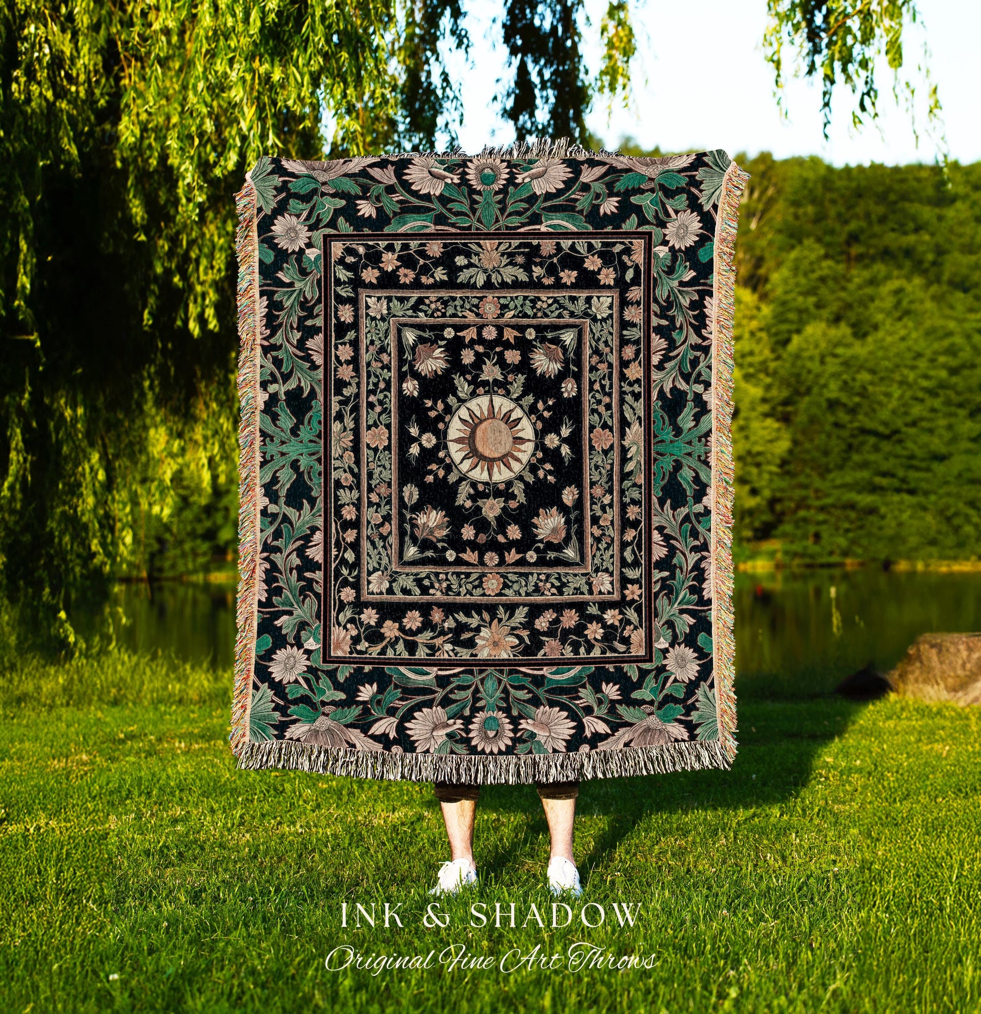 May include: A woven tapestry throw blanket with a black background and a gold sun in the center. The sun is surrounded by pink and green floral designs. The blanket has a fringe border and is made of a soft, luxurious fabric. The text &#39;INK & SHADOW Original Fine Art Throws&#39; is printed below the blanket.