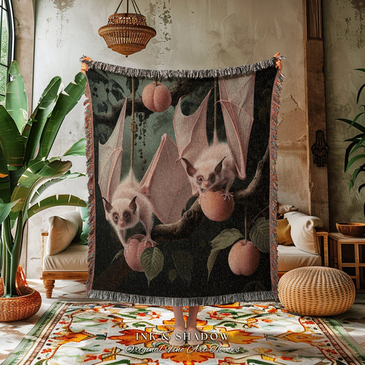 May include: A woven tapestry throw blanket with a dark green background featuring two pink bats with white fur. The bats are hanging from branches with pink and orange fruit. The tapestry has a fringe border and the words &#39;INK & SHADOW Original Fine Art Throws&#39; are printed at the bottom.