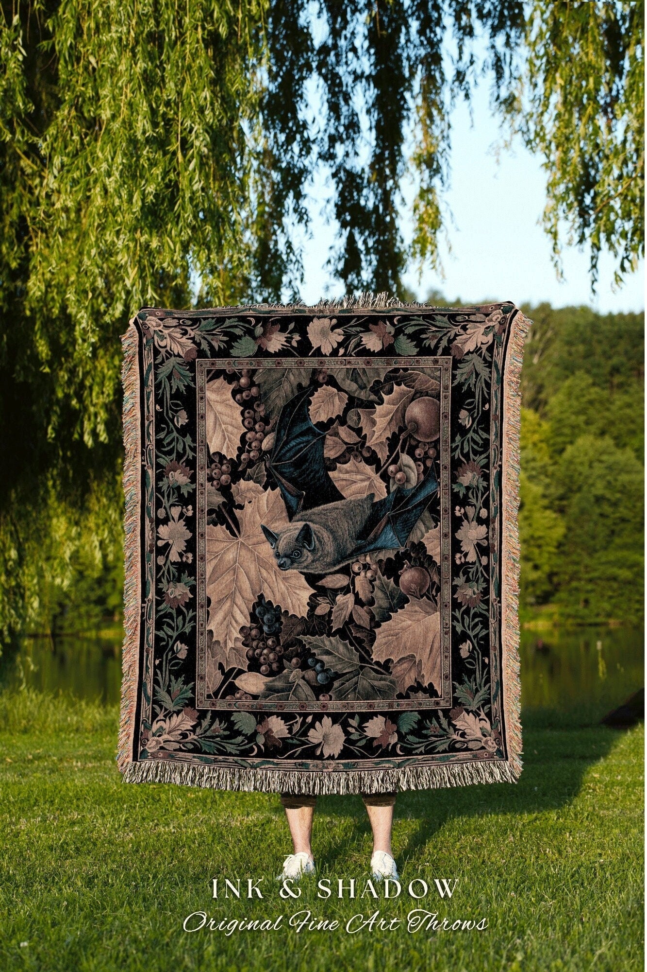 May include: A woven tapestry throw blanket with a black background and a detailed design of a bat flying through a vine of grapes. The throw blanket has a fringe border and the text &#39;INK & SHADOW Original Fine Art Throws&#39; is displayed below the blanket.