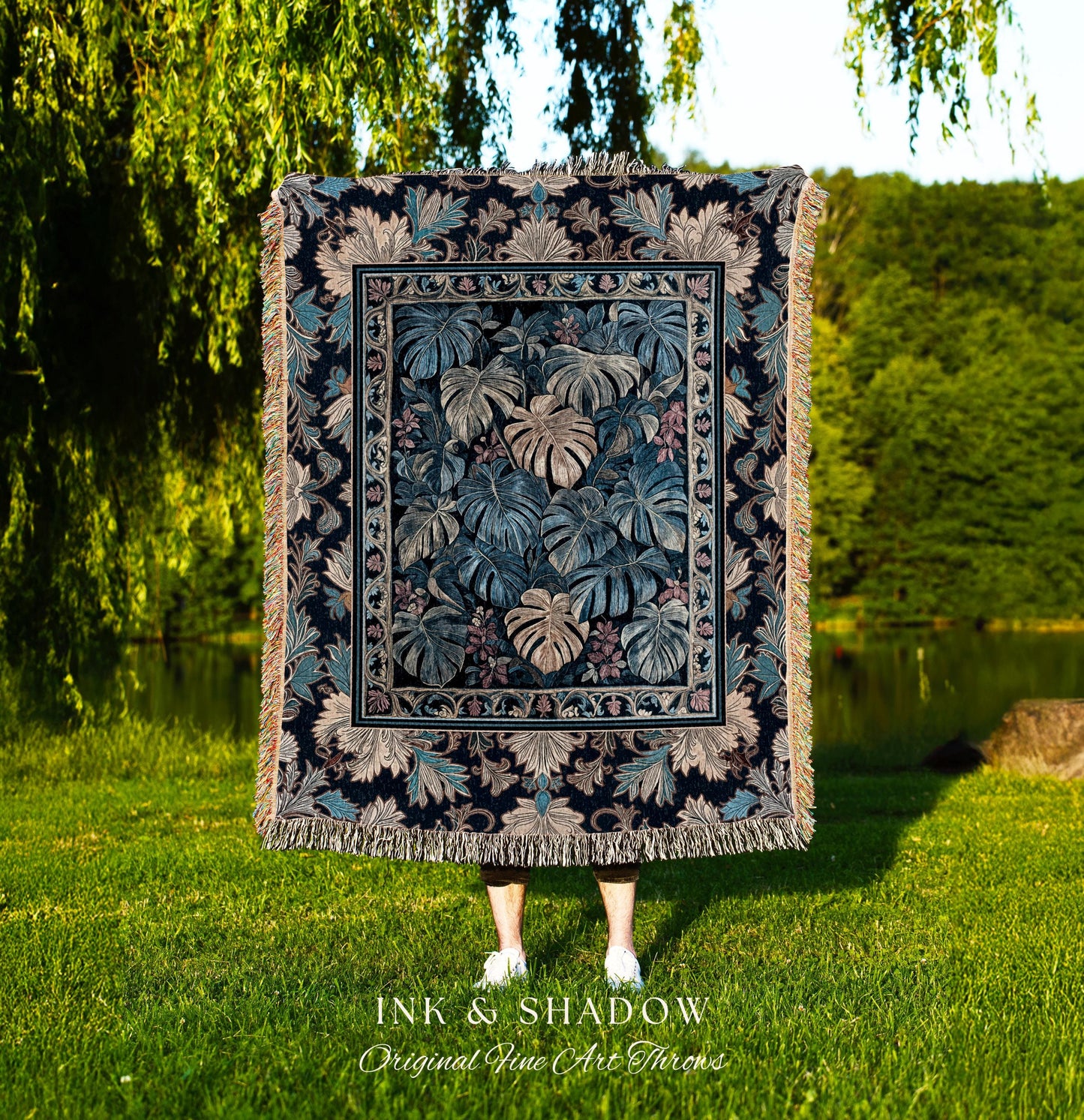 May include: A woven tapestry throw blanket with a dark blue background and a pattern of large green and beige leaves. The throw blanket is fringed on all sides. The text &#39;INK & SHADOW Original Fine Art Throws&#39; is printed below the blanket.