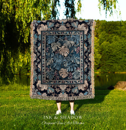May include: A woven tapestry throw blanket with a dark blue background and a pattern of large green and beige leaves. The throw blanket is fringed on all sides. The text &#39;INK & SHADOW Original Fine Art Throws&#39; is printed below the blanket.