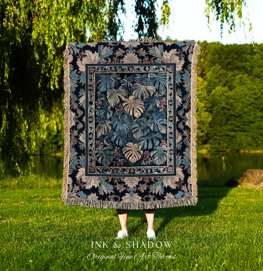 May include: A woven tapestry throw blanket with a dark blue background and a pattern of large green and beige leaves. The throw blanket is fringed on all sides. The text &#39;INK & SHADOW Original Fine Art Throws&#39; is printed below the blanket.