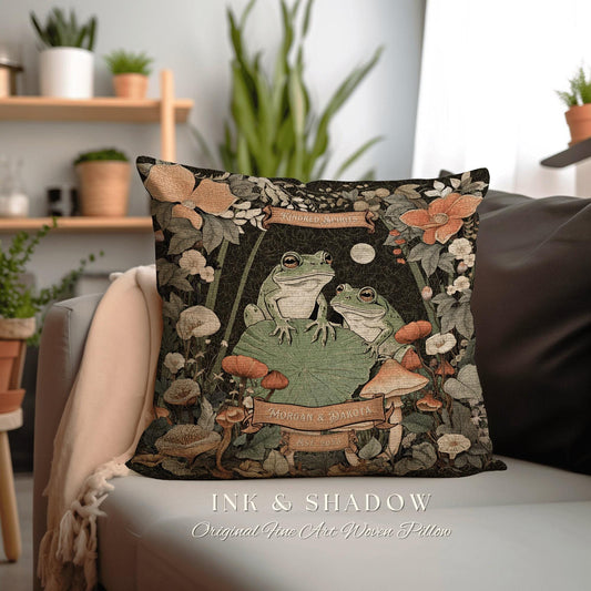 May include: A woven pillow with a dark green background featuring two green frogs sitting on a lily pad. The frogs are surrounded by flowers, mushrooms, and vines. The pillow has a banner that reads 'Kindred Spirits' and another banner that reads 'Morgan & Dakota Est. 2023'. The pillow is labeled 'Ink & Shadow Original Fine Art Woven Pillow'.