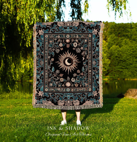 May include: A woven tapestry throw blanket with a black background and a gold sun and moon design. The sun and moon are surrounded by a floral pattern in shades of blue and green. The blanket has fringe on all four sides. The text 'INK & SHADOW Original Fine Art Throws' is printed below the blanket.