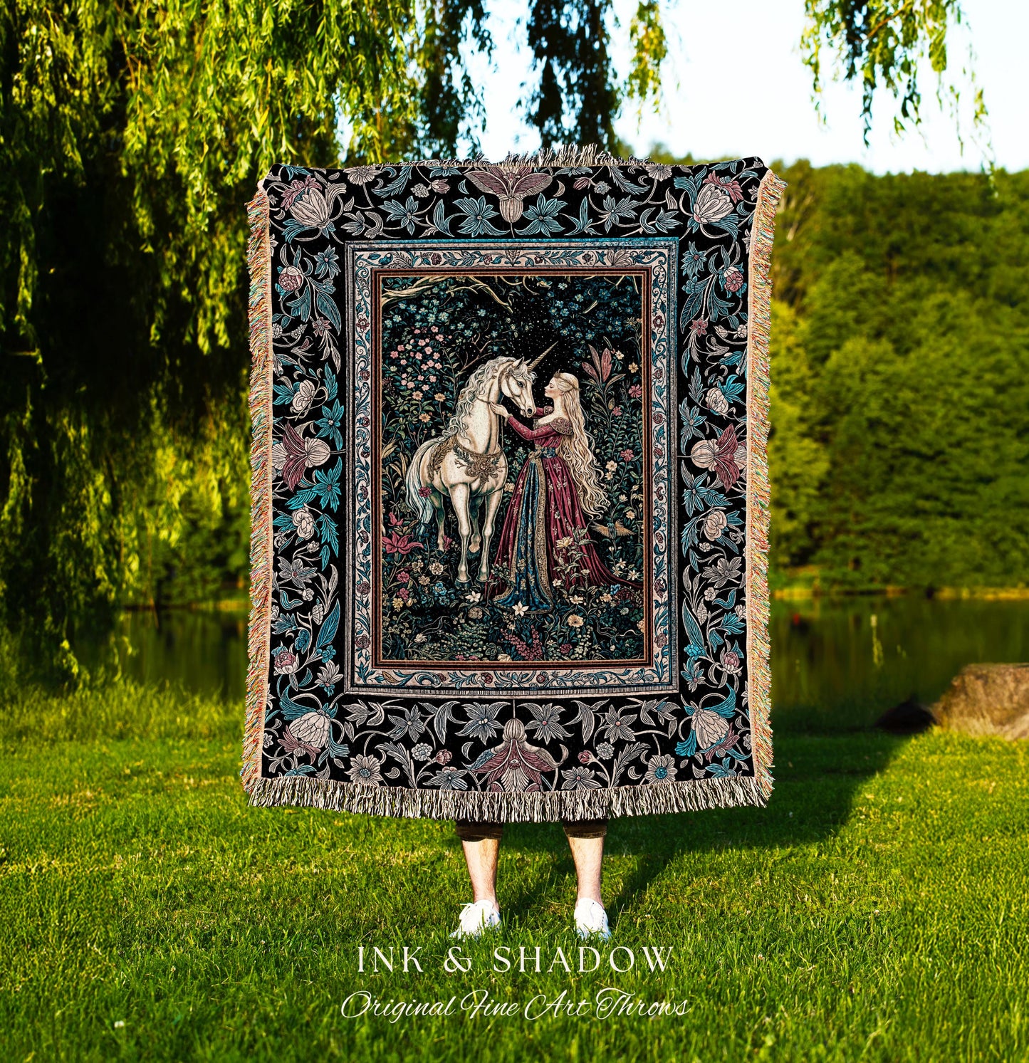 May include: A woven tapestry throw blanket with a black background and a colorful floral border. The center of the blanket features a woman in a long red dress with long blonde hair, kneeling and touching her hand to the head of a white unicorn. The text 'INK & SHADOW Original Fine Art Throws' is printed below the blanket.
