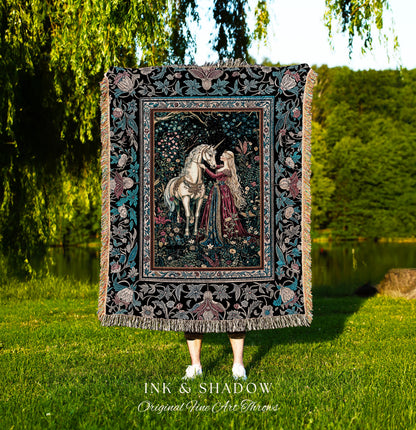 May include: A woven tapestry throw blanket with a black background and a colorful floral border. The center of the blanket features a woman in a long red dress with long blonde hair, kneeling and touching her hand to the head of a white unicorn. The text 'INK & SHADOW Original Fine Art Throws' is printed below the blanket.