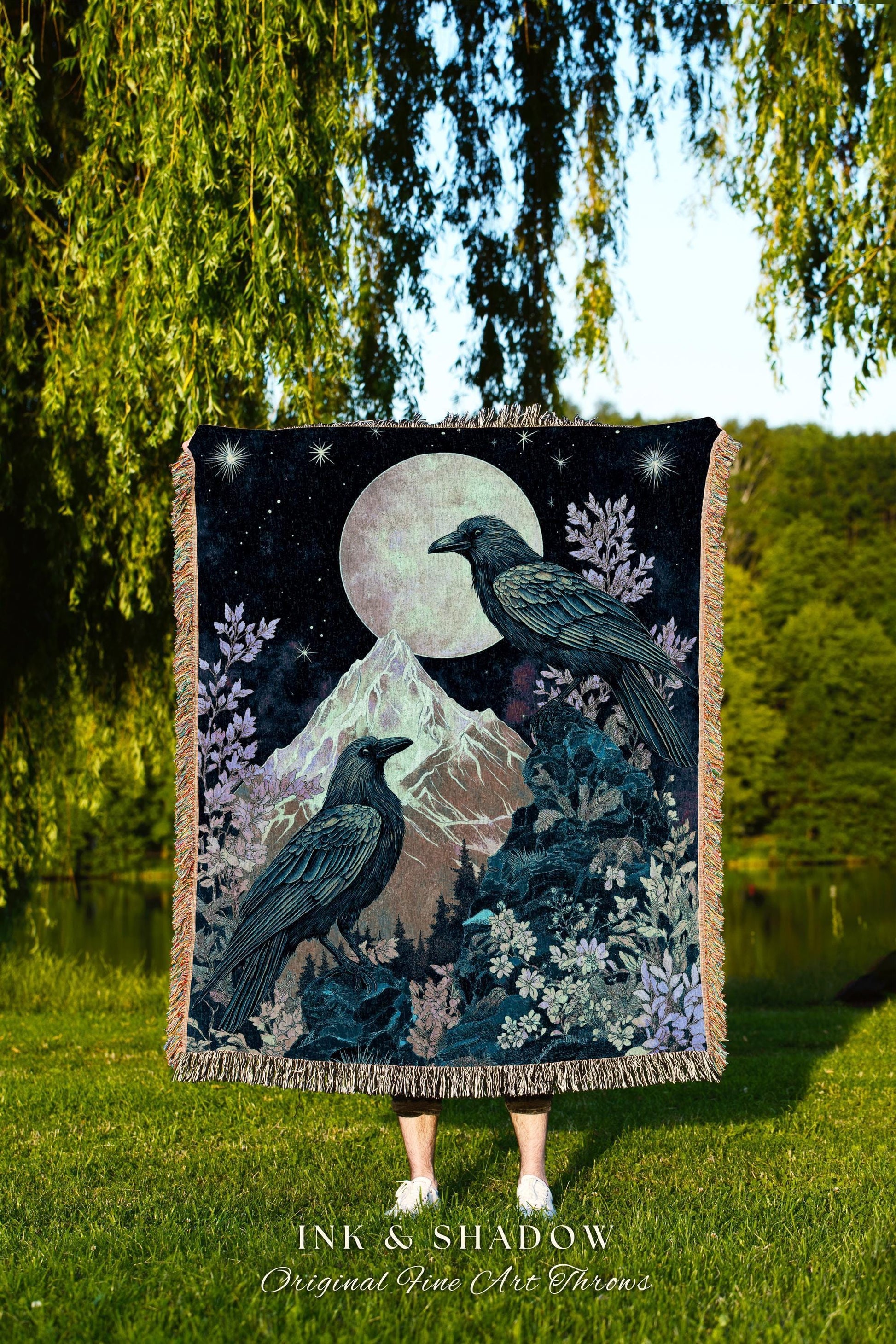 May include: A woven tapestry throw blanket with a dark blue background. The blanket features two black crows perched on a mountain with a full moon in the background. The moon is a light blue color. The blanket has a fringe border and the words 'INK & SHADOW Original Fine Art Throws' are printed at the bottom.