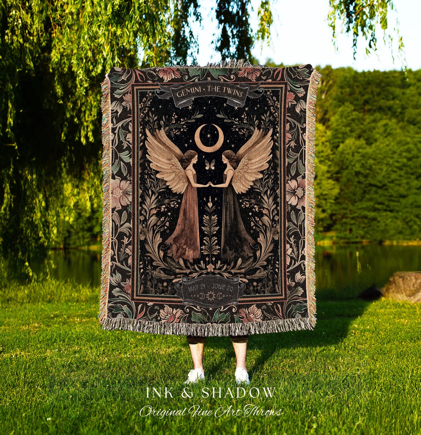 May include: A woven tapestry throw blanket with a dark background and floral accents. The center of the blanket features a black and white illustration of two women with wings, a crescent moon, and the text 'GEMINI THE TWINS' and 'MAY 21 - JUNE 20'. The bottom of the blanket has the text 'INK & SHADOW Original Fine Art Throws'.