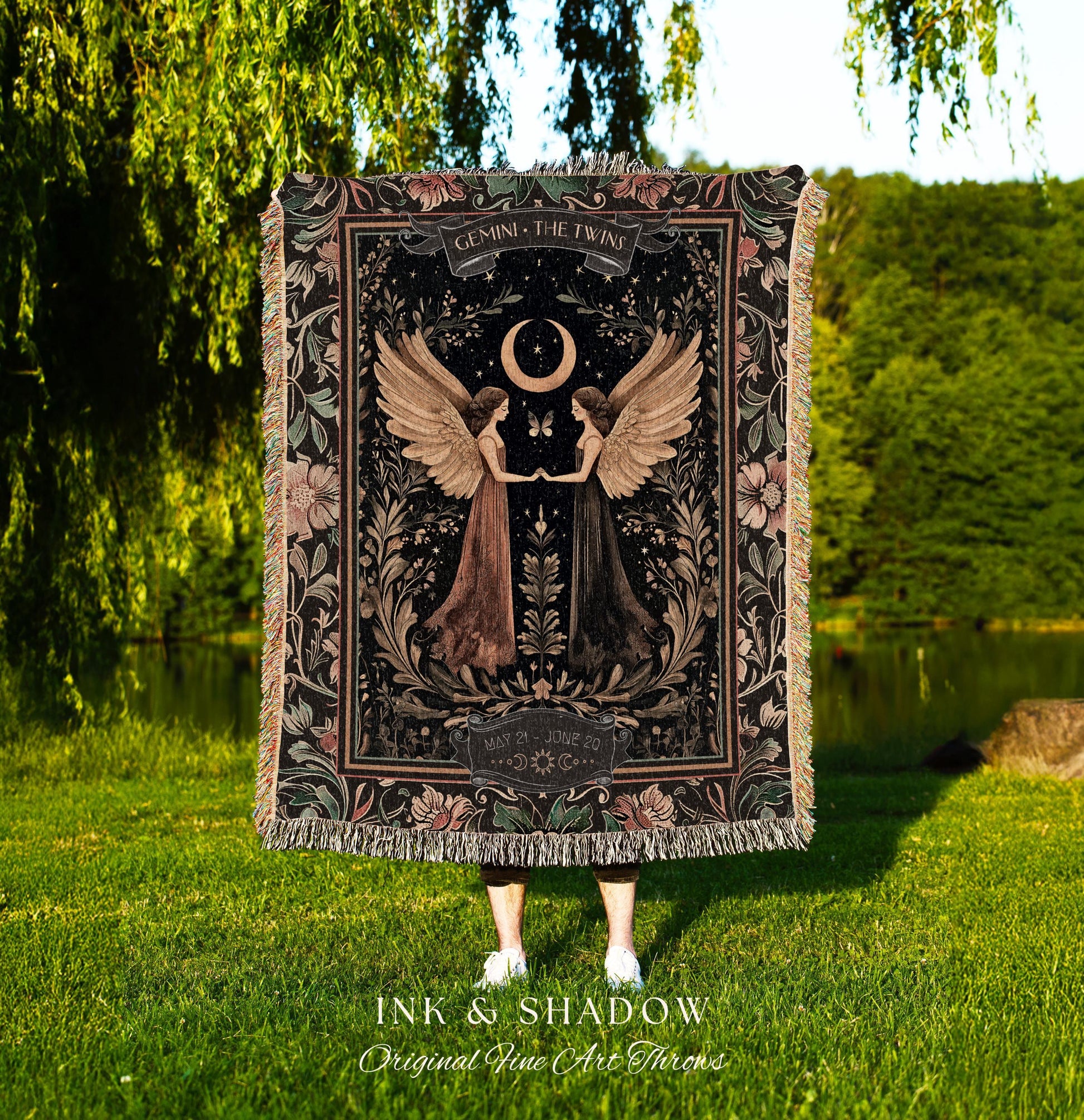 May include: A woven tapestry throw blanket with a dark background and floral accents. The center of the blanket features a black and white illustration of two women with wings, a crescent moon, and the text 'GEMINI THE TWINS' and 'MAY 21 - JUNE 20'. The bottom of the blanket has the text 'INK & SHADOW Original Fine Art Throws'.
