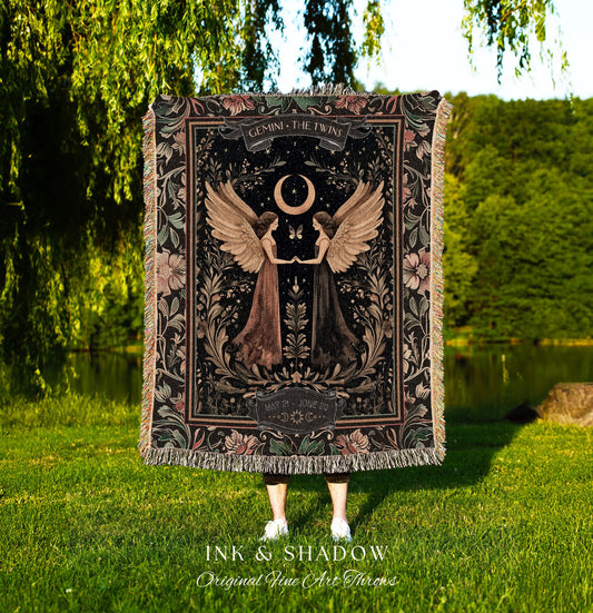 May include: A woven tapestry throw blanket with a dark background and floral accents. The center of the blanket features a black and white illustration of two women with wings, a crescent moon, and the text 'GEMINI THE TWINS' and 'MAY 21 - JUNE 20'. The bottom of the blanket has the text 'INK & SHADOW Original Fine Art Throws'.