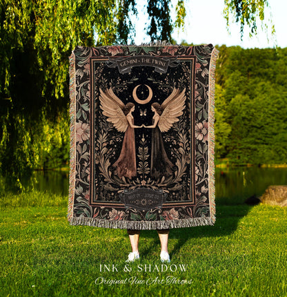May include: A woven tapestry throw blanket with a dark background and floral accents. The center of the blanket features a black and white illustration of two women with wings, a crescent moon, and the text 'GEMINI THE TWINS' and 'MAY 21 - JUNE 20'. The bottom of the blanket has the text 'INK & SHADOW Original Fine Art Throws'.