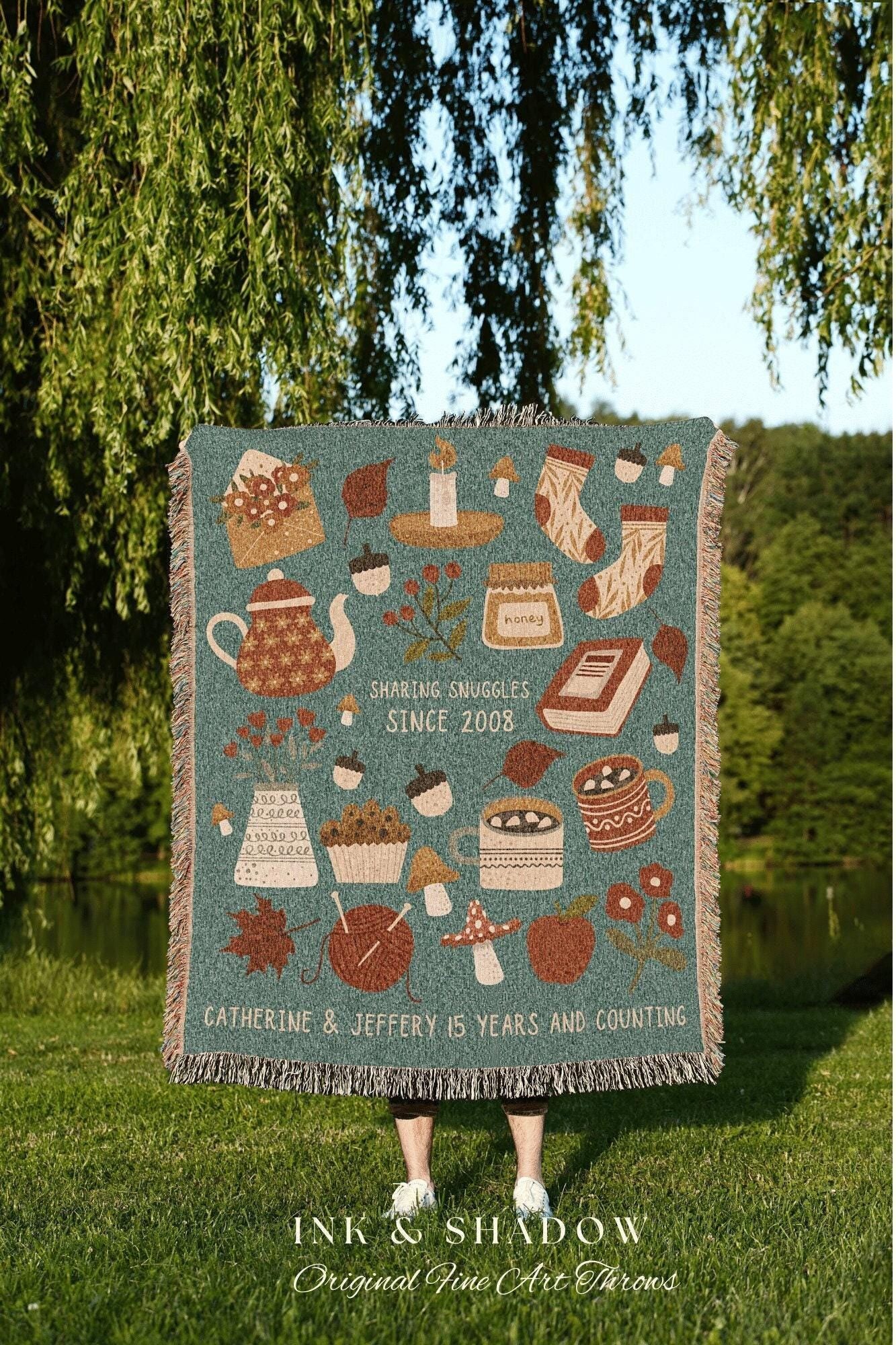 May include: A woven throw blanket with a teal background and a variety of autumn-themed illustrations, including a teapot, a candle, a jar of honey, a book, a cupcake, mushrooms, a mug of hot chocolate, and a ball of yarn. The text on the blanket reads 'Sharing Snuggles Since 2008' and 'Catherine & Jeffery 15 Years and Counting.' The blanket is made by Ink & Shadow Original Fine Art Throws.